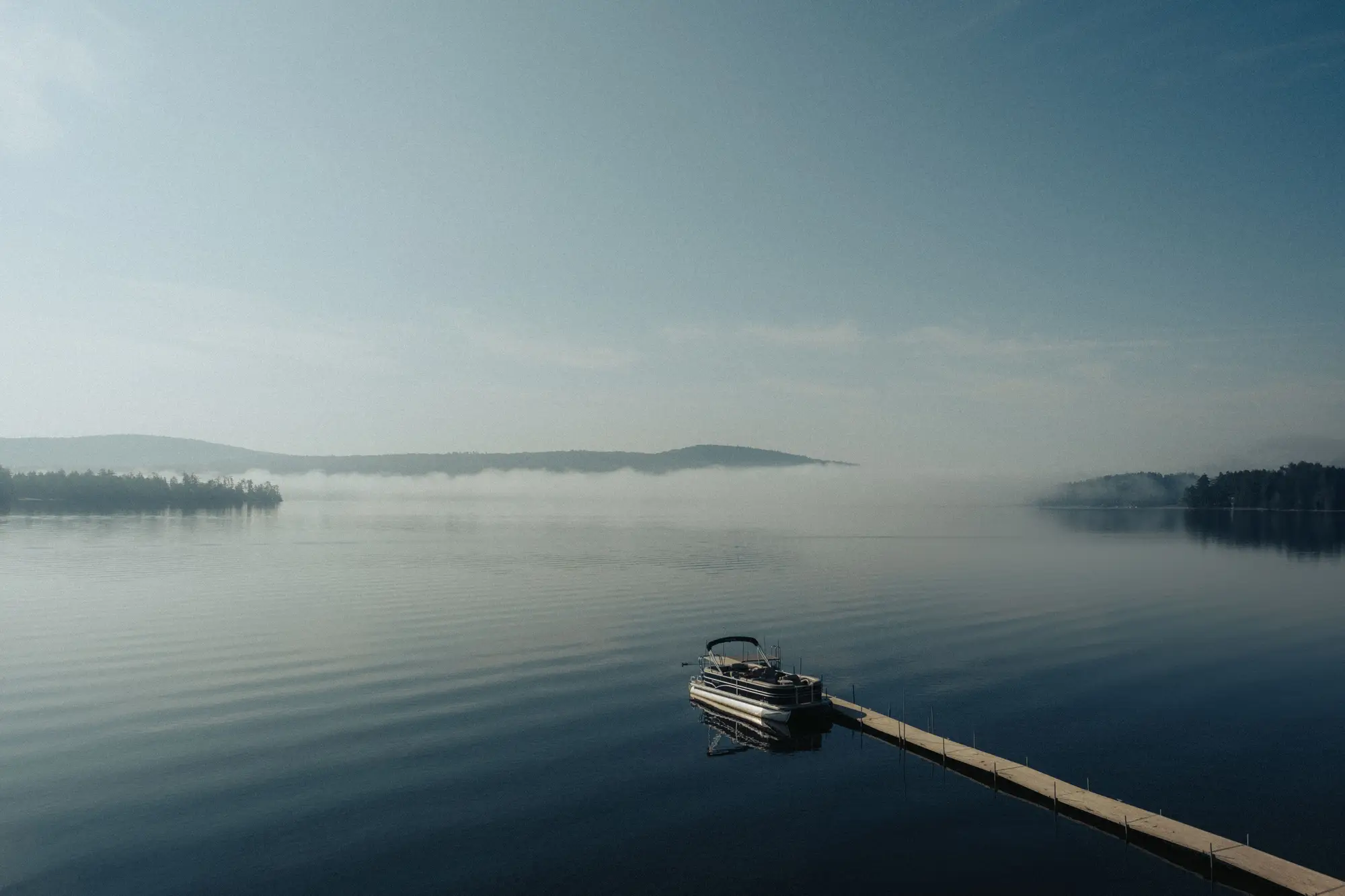 A pontoon boat tied up at the end of a long dock on a calm lake, mist lying over the far shore
