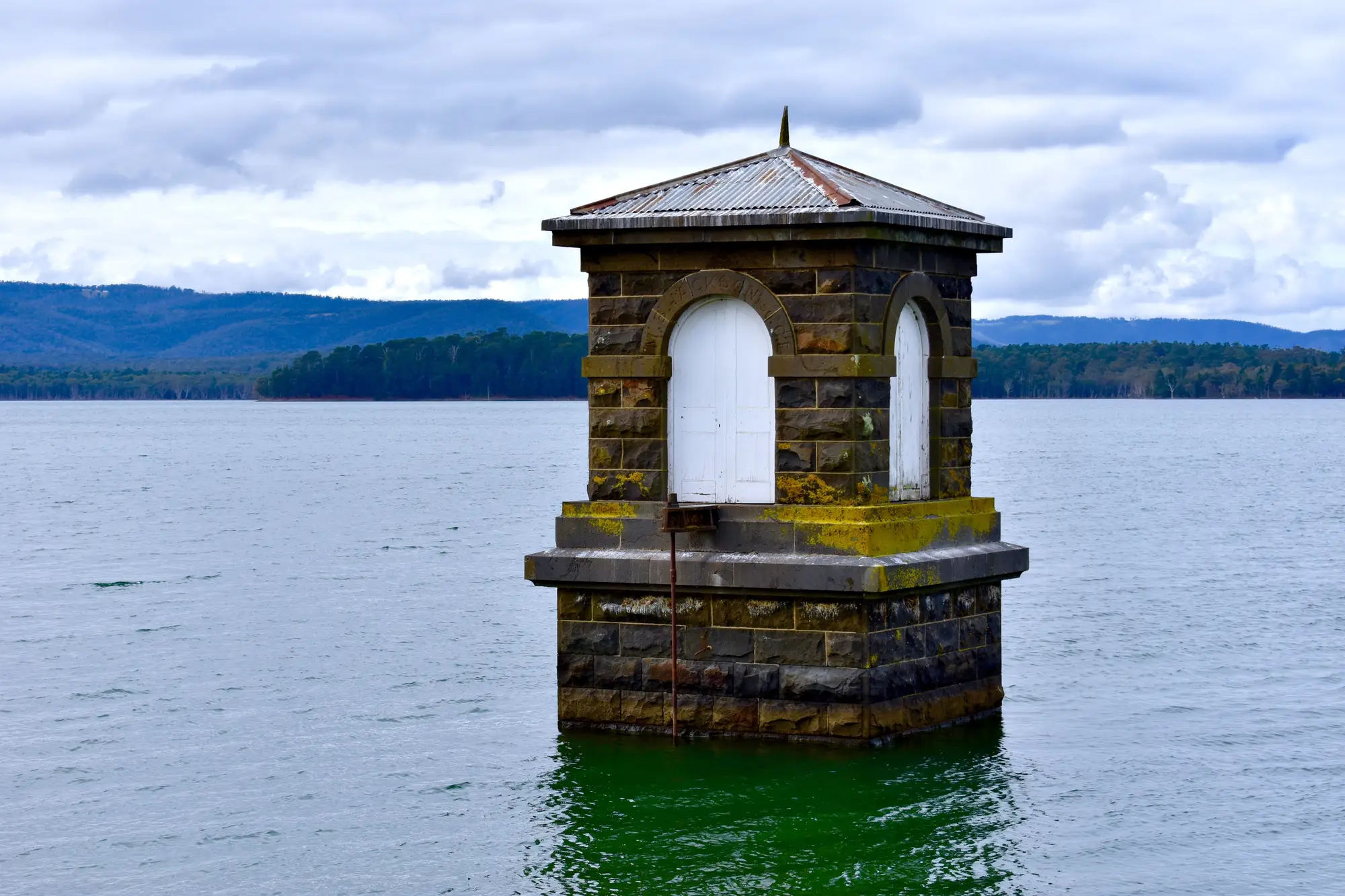 A stone valve tower standing in a reservoir, green water at its base and wooded hills behind